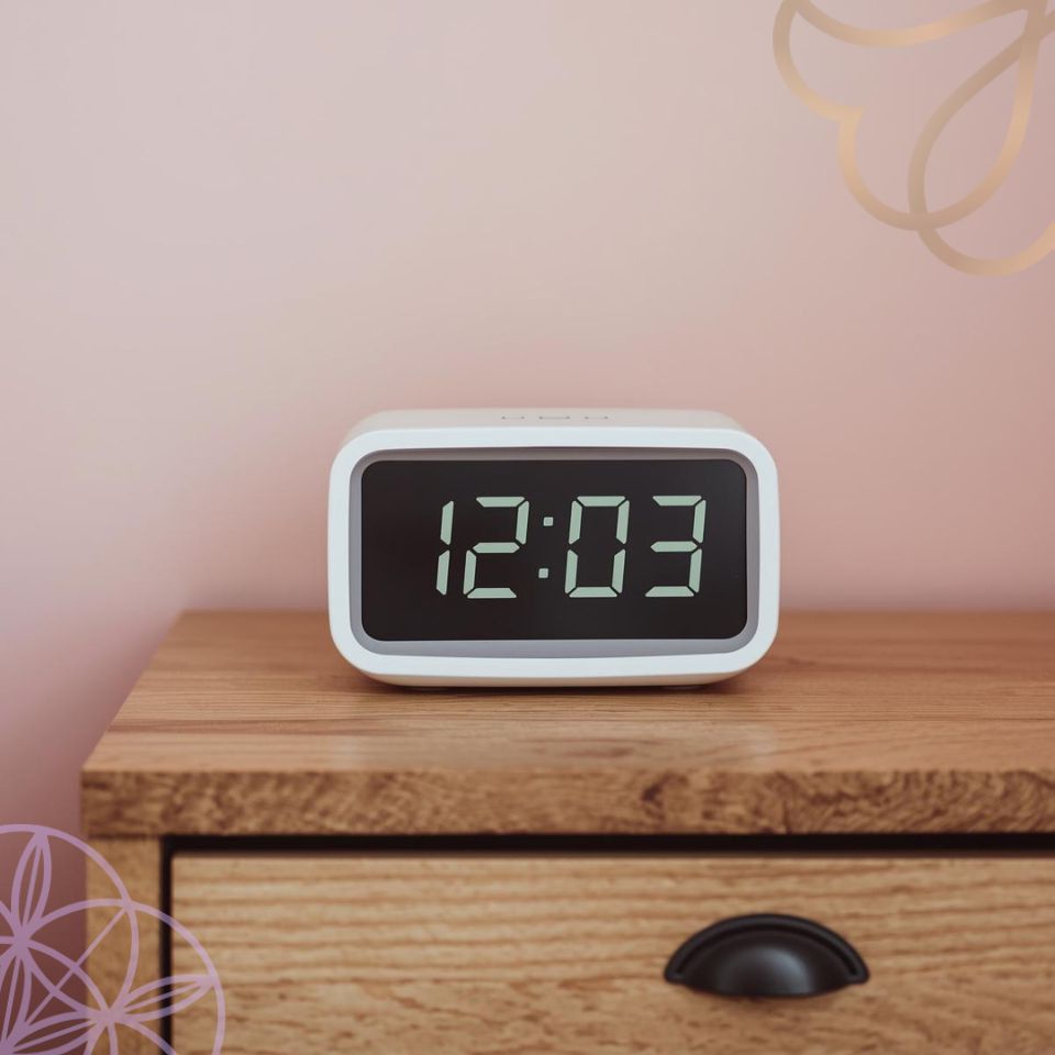 a close-up of a white digital clock that reads 12:03. It should be on top of a jarrah wooden bedside table with pastel pink wall behind it