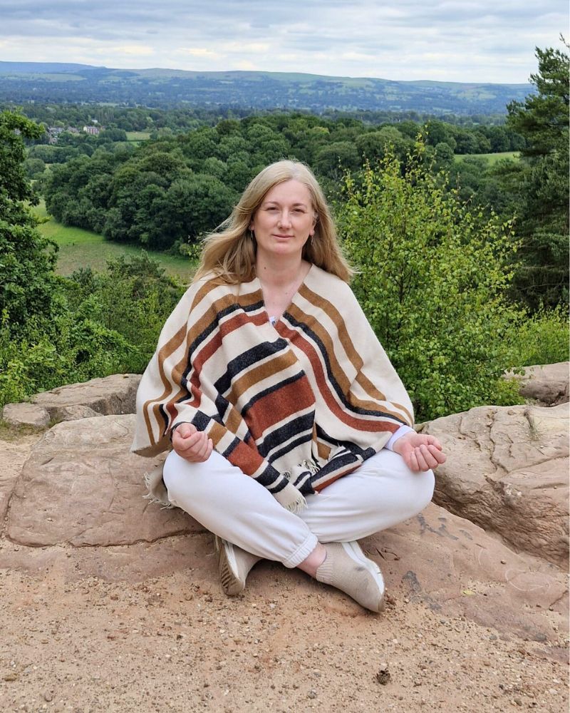 Johanna sitting cross-legged on a rock overlooking a forest landscape, representing calm, presence and grounded awareness