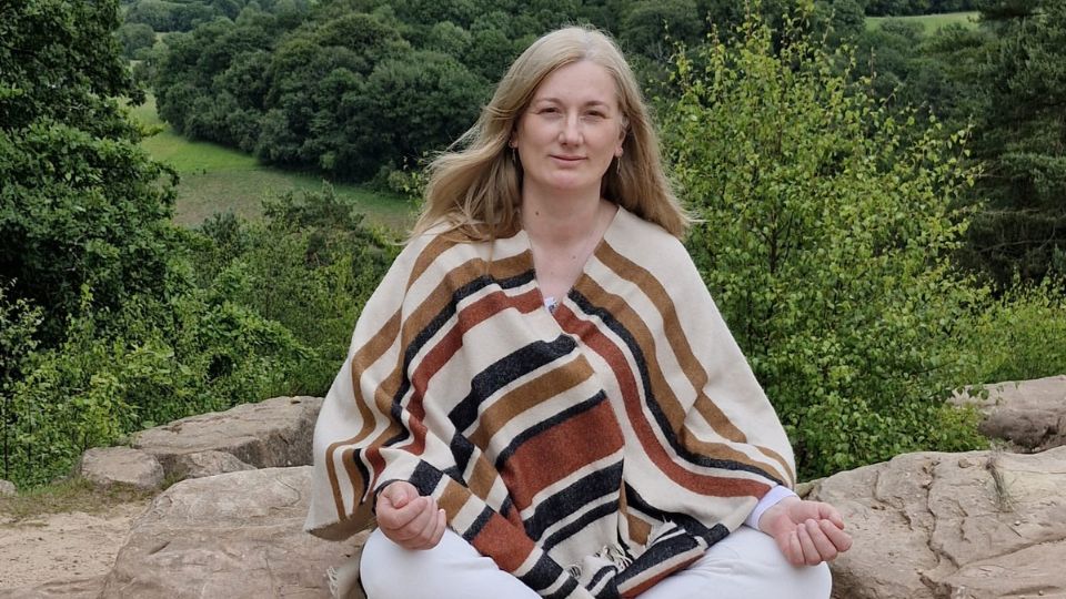 Johanna sitting cross-legged on a rock overlooking a forest landscape, representing calm, presence and grounded awareness