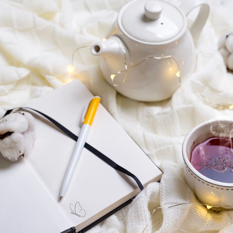 Open journal with pen beside a teapot and cup of tea on a soft blanket, representing reflection and guided journaling prompts