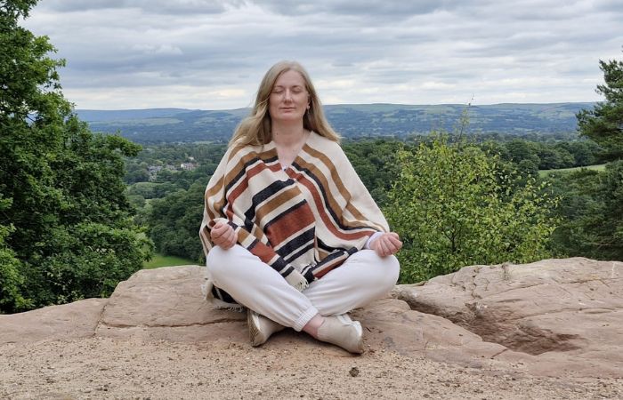 SOIE-soul-aligned-meditation Johanna sitting cross-legged with her eyes closed in meditation on a rocky lookout, overlooking a wide green valley landscape