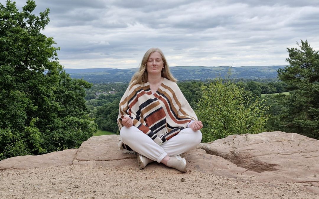 Soie-2024-born Johanna sitting cross-legged on a rock overlooking a forest landscape with eyes closed, representing stillness and inner awareness